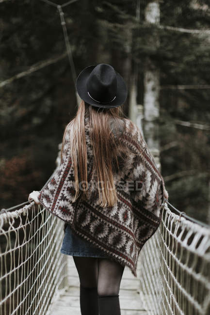 Back view of fashionable young woman wearing hat and poncho and walking on suspension bridge — Stock Photo