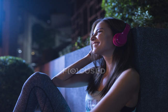 Mujer joven feliz con auriculares rosados escuchando música en un entorno urbano moderno por la noche - foto de stock