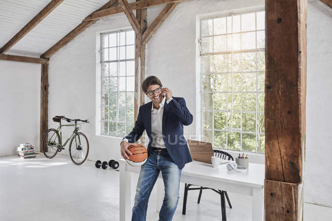 Hombre de negocios sonriente con baloncesto en el teléfono celular en el ático - foto de stock