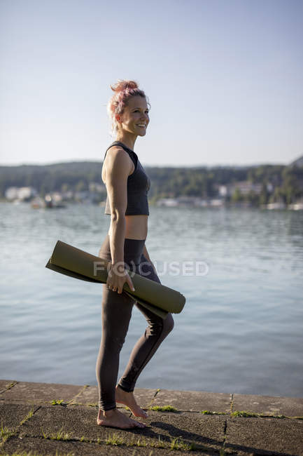 Женщина в спортивной одежде ходит с ковриком для йоги на берегу озера — стоковое фото