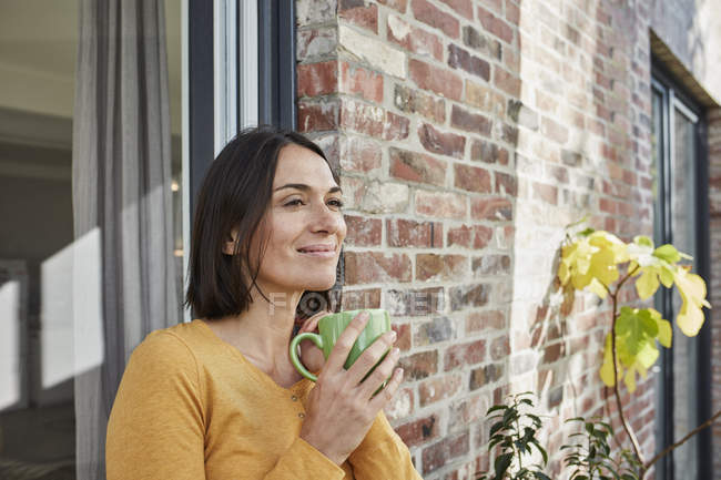 Mujer sonriente bebiendo de la taza frente a su casa - foto de stock