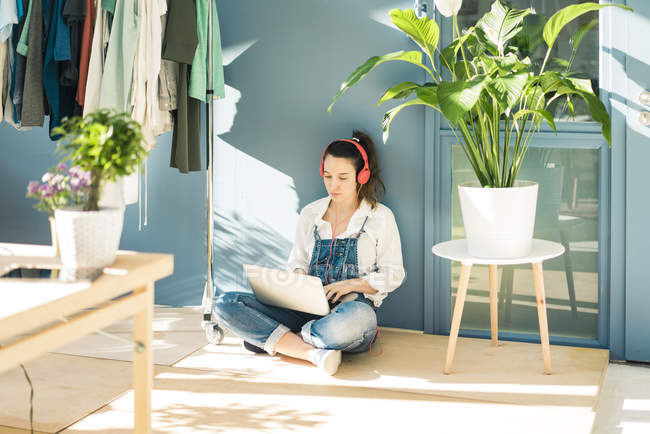 Joven freelancer sentada en el suelo en su estudio usando laptop y auriculares - foto de stock