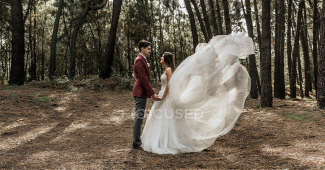 Noiva com vestido de noiva varrido pelo vento e noivo de pé na floresta de mãos dadas — Fotografia de Stock