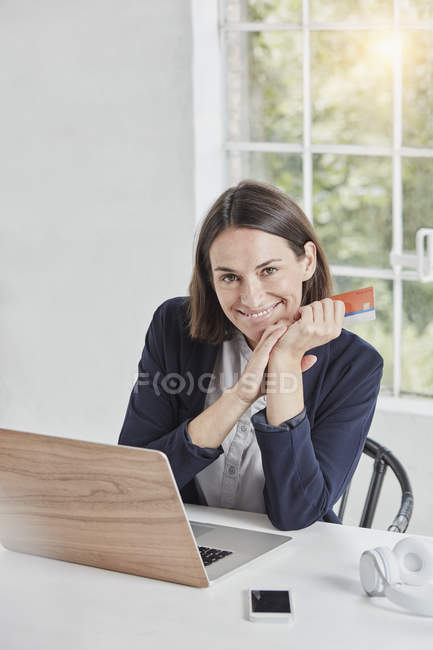 Portrait of smiling businesswoman with laptop on desk holding card — Stock Photo