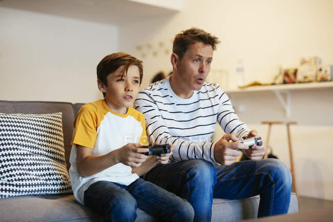 Padre e hijo jugando videojuegos en el sofá en casa - foto de stock
