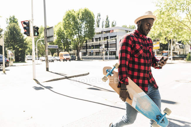 Cooler junger Mann läuft mit Skateboard durch die Stadt und nutzt Smartphone — Stockfoto