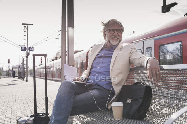 Sorridente uomo d'affari maturo seduto alla stazione ferroviaria con cellulare, auricolari e notebook — Foto stock