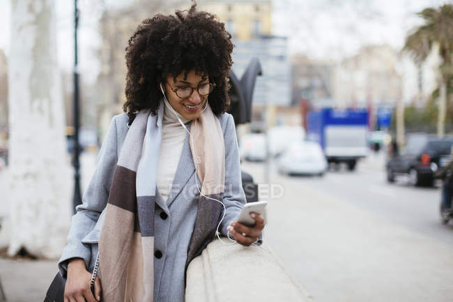 España, Barcelona, mujer sonriente con teléfono celular y auriculares caminando por la ciudad - foto de stock