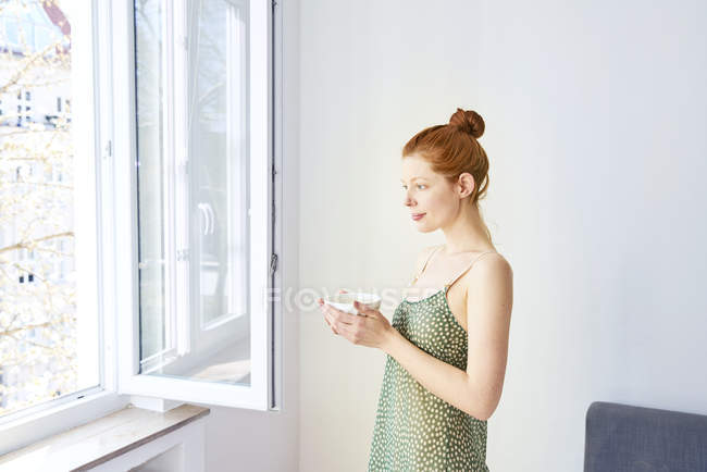 Портрет задумчивой женщины с чашкой белого кофе, стоящей у открытого окна — стоковое фото