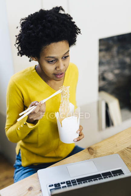 Mujer afroamericana joven sentada en la mesa, navegar por la web en el ordenador portátil mientras come fideos - foto de stock