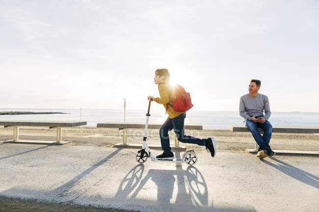 Vater beobachtet Sohn beim Rollerfahren auf Strandpromenade bei Sonnenuntergang — Stockfoto