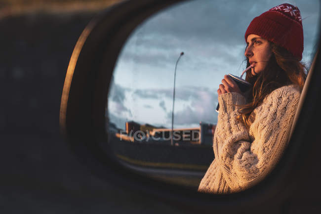 Iceland, young woman with coffee to go at sunset, mirrored in wing mirror — Stock Photo