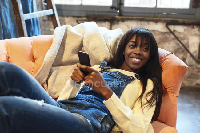 Retrato de una mujer sonriente acostada en el sofá de su casa usando un teléfono celular - foto de stock