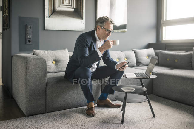 Mature businessman with cup of coffee and laptop using cell phone on couch — Stock Photo
