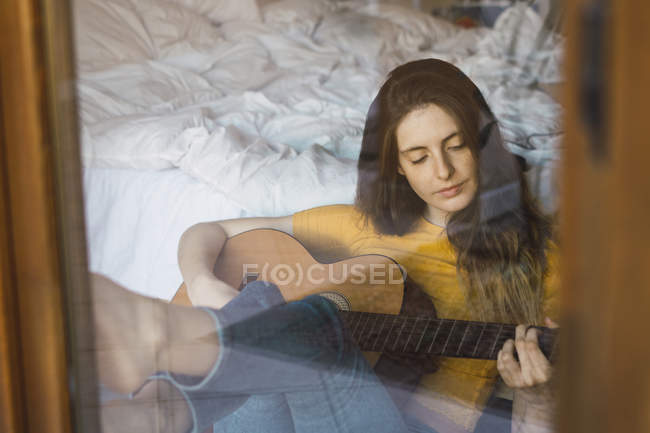Retrato de una joven relajada sentada detrás del cristal de la ventana tocando la guitarra - foto de stock