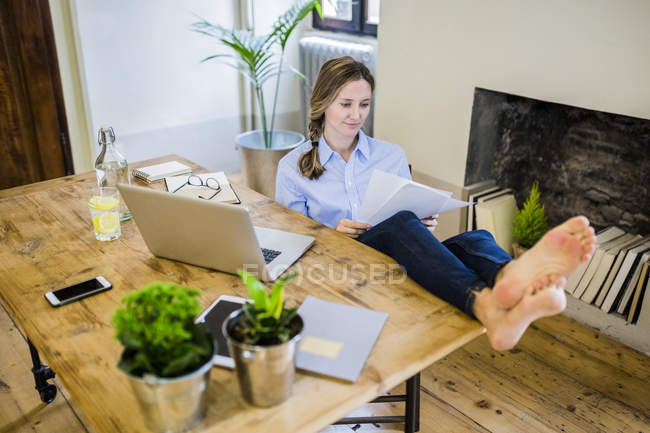 Женщина сидит за столом дома с поднятыми ногами и читает документ — стоковое фото