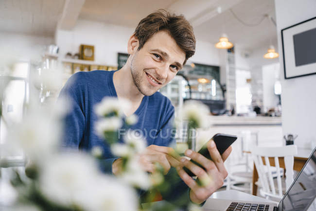 Ritratto di uomo sorridente in un caffè con cellulare e laptop — Foto stock