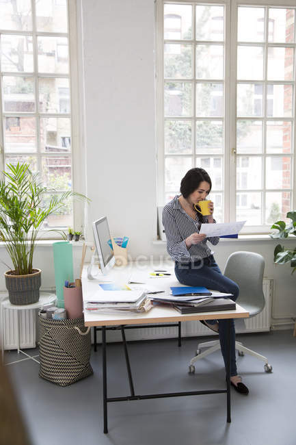 Frau mit Tasse Kaffee blickt auf Papier am Schreibtisch im Büro — Stockfoto