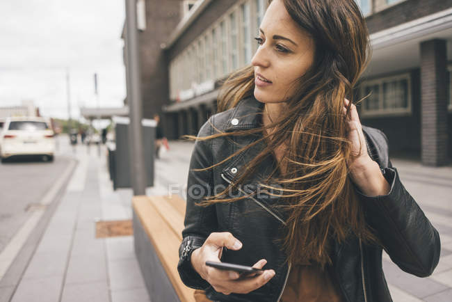Giovane donna con i capelli spazzati dal vento che tiene il telefono cellulare in città — Foto stock