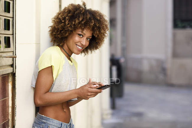 Портрет улыбающейся женщины с африканской прической с использованием смартфона на открытом воздухе — стоковое фото