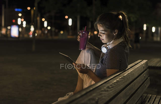 Mujer joven con batido sentado en el banco por la noche usando la tableta - foto de stock