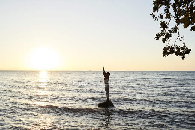 Jeune femme pratiquant le yoga sur un rocher dans la mer au coucher du soleil — Photo de stock