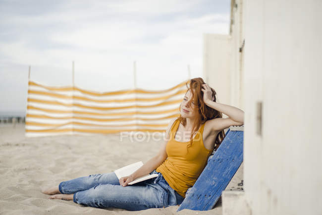 Donna rossa seduta davanti alla cabina sulla spiaggia, che legge un libro — Foto stock