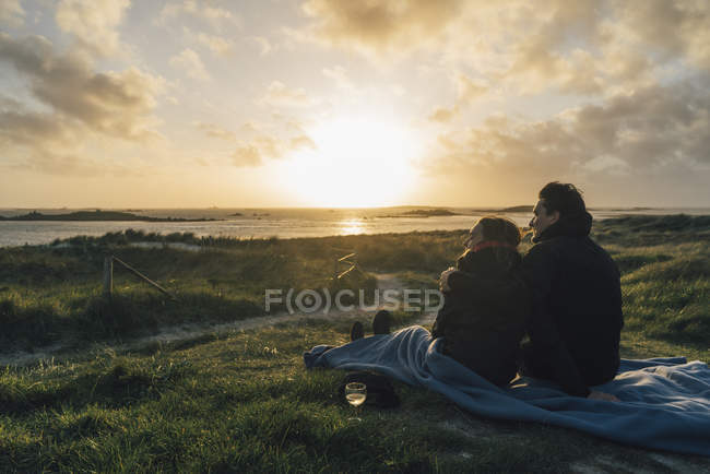 Francia, Bretaña, Landeda, pareja sentada en la costa al atardecer - foto de stock
