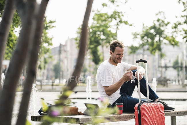 Homme souriant avec valise roulante assise sur un banc — Photo de stock