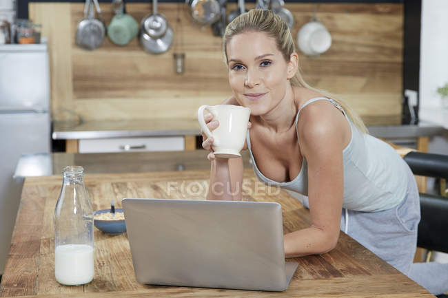 Retrato de mulher loira sorridente com laptop na cozinha bebendo café — Fotografia de Stock