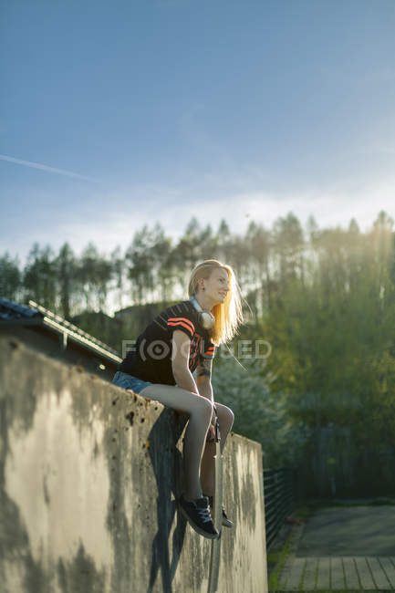 Улыбающаяся блондинка с длинной доской сидит на стене и смотрит вдаль — стоковое фото