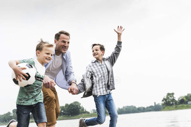 Pai feliz com dois filhos e futebol correndo à beira do rio — Fotografia de Stock
