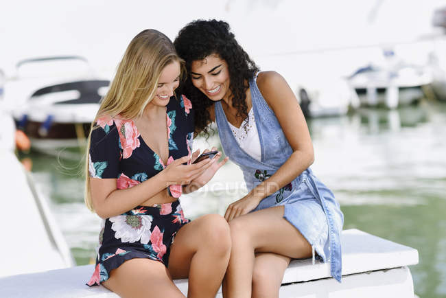 Due giovani donne felici con il cellulare al porto turistico in estate — Foto stock