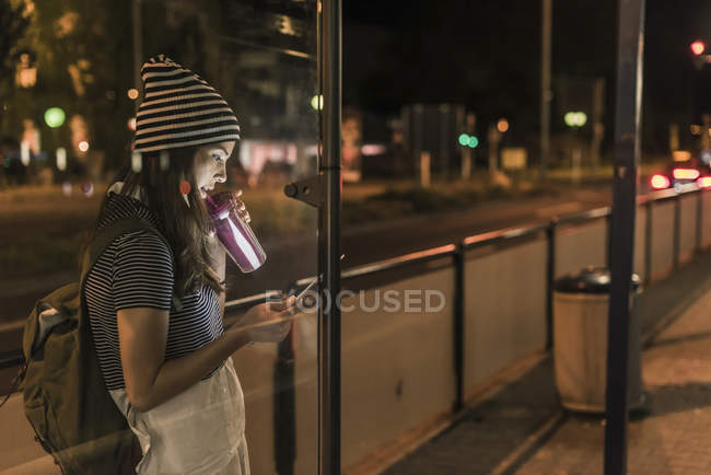 Jovem com smoothie esperando na estação à noite olhando para o telefone celular — Fotografia de Stock