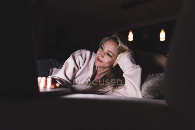 Smiling woman lying on couch at home using laptop — Stock Photo