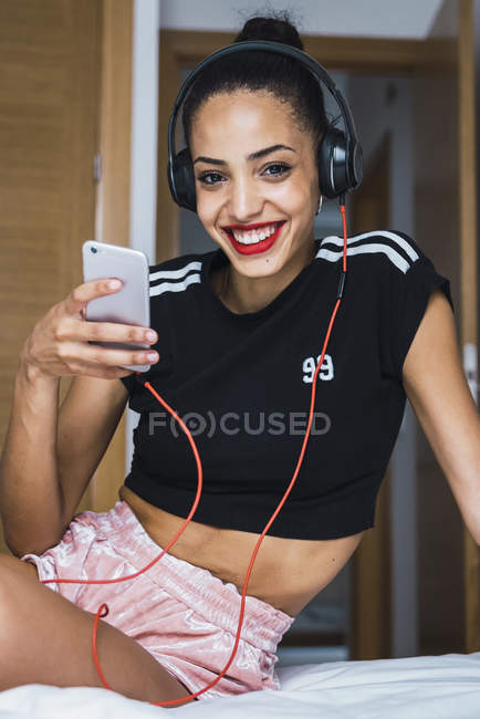 Портрет усмішки прекрасної молодої жінки, яка сидить на ліжку з мобільним телефоном і навушниками. — стокове фото
