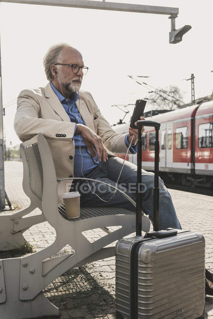 Uomo d'affari maturo seduto alla stazione ferroviaria con cellulare, auricolari e valigia — Foto stock