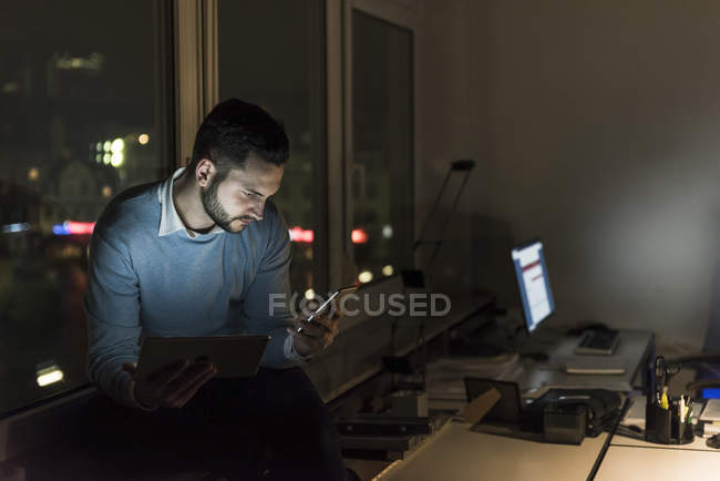 Бізнесмен сидить на підвіконні в офісі вночі, дивлячись на смартфон — стокове фото