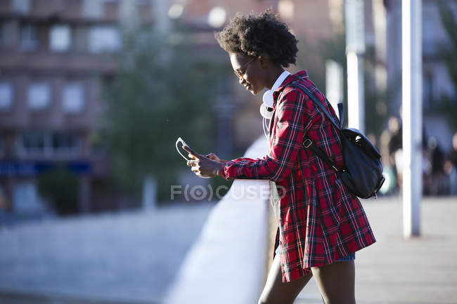 Молода жінка з навушниками і рюкзаком стоїть на мосту за допомогою мобільного телефону. — стокове фото