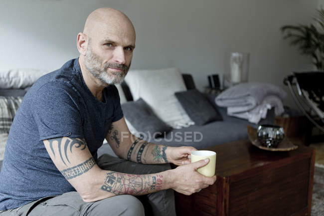 Татуйований чоловік вдома сидить на дивані, п'є каву — стокове фото