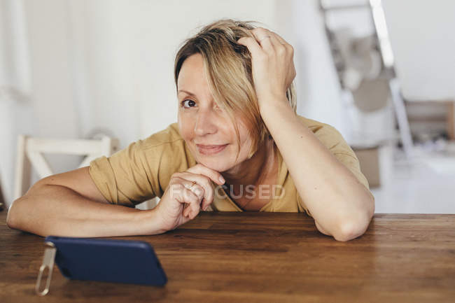 Porträt einer lächelnden Frau, die mit Smartphone in neuer Wohnung am Tisch sitzt — Stockfoto