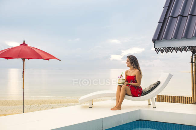 Thailandia, Koh Phangan, donna felice seduta a bordo piscina vicino alla spiaggia guardando la distanza — Foto stock