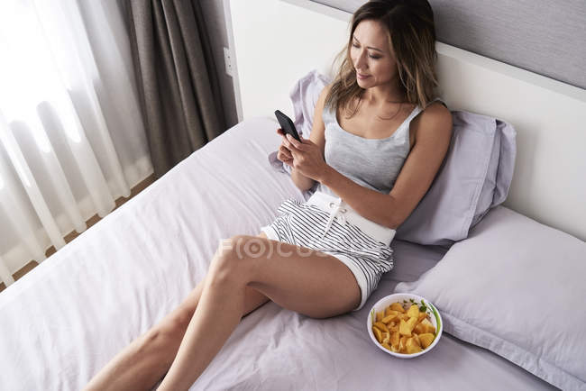 Beautiful woman using smartphone in bed while breakfasting a healthy fruit bowl — Stock Photo
