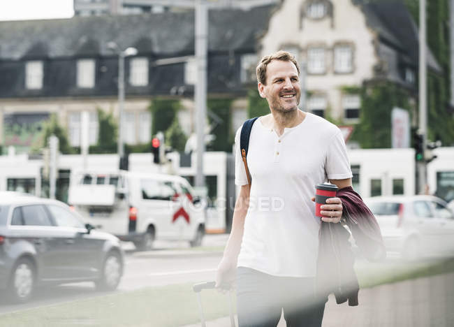 Улыбающийся мужчина с чемоданом на колесах и кофе на вынос гуляет по городу — стоковое фото