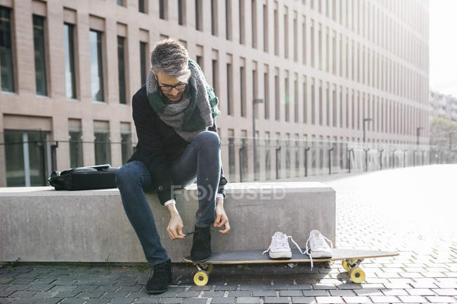 Freelancer con longboard sentado en el banco y atándose los zapatos - foto de stock