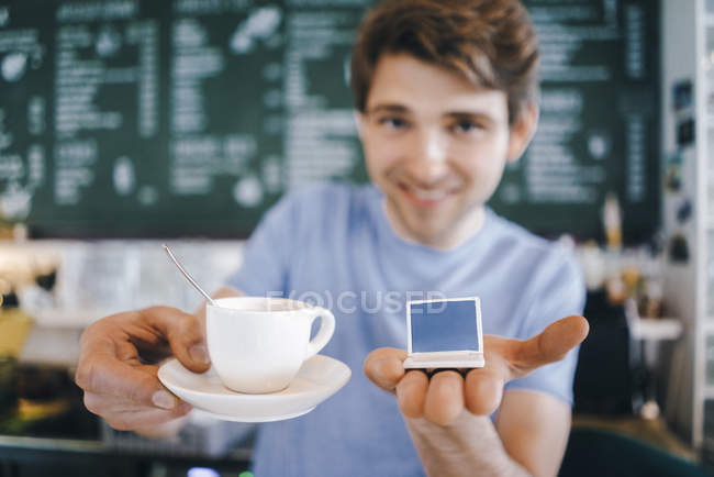 Усміхнений чоловік в кафе пропонує чашку кави і тримає мініатюрну модель ноутбука — стокове фото