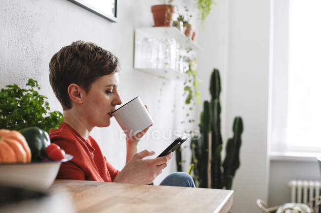 Женщина сидит на кухне, пьет кофе и проверяет сообщения со смартфона — стоковое фото