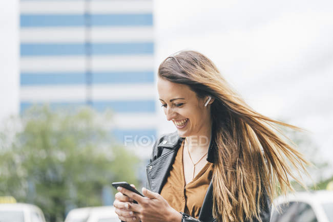 Joyeux jeune femme avec les cheveux balayés par le vent en utilisant un téléphone portable dans la ville — Photo de stock