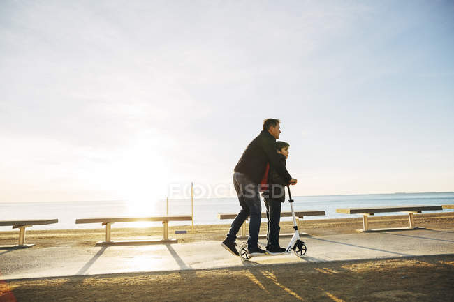 Батько і син їзда на скутері на пляжній набережній на заході сонця — стокове фото