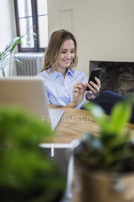 Donna sorridente seduta alla scrivania a casa e controllare il telefono cellulare — Foto stock